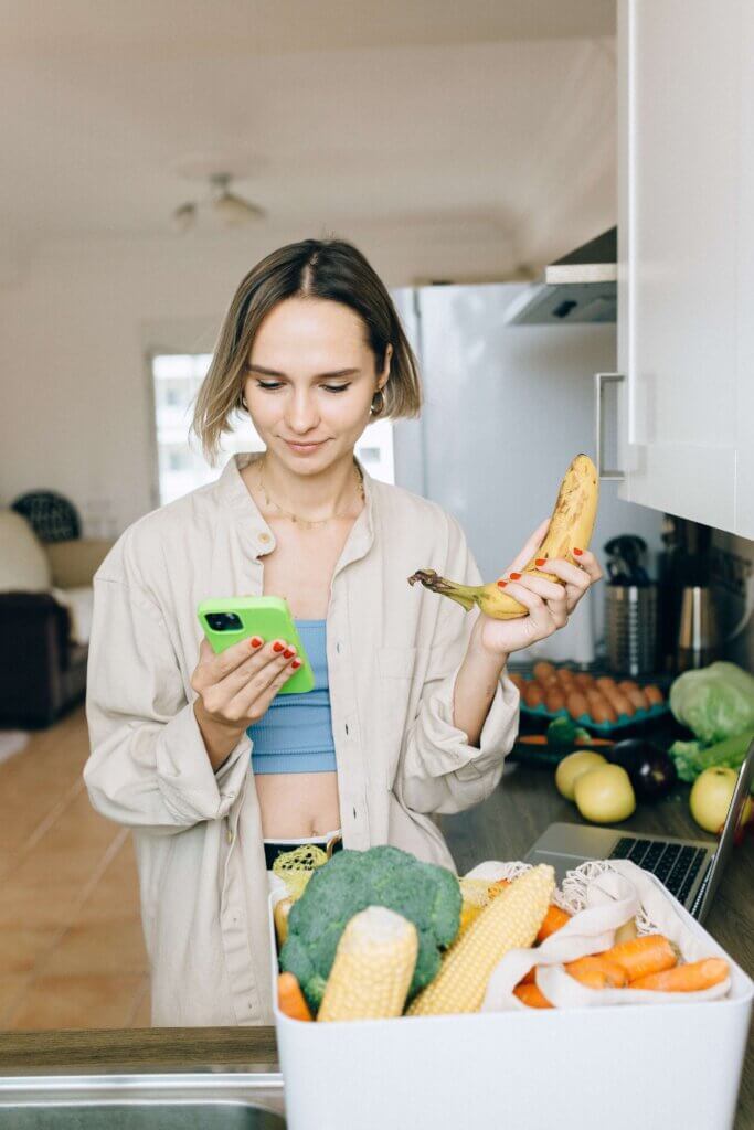 Woman holding a banana and looking at her phone with vegetables out on the counter